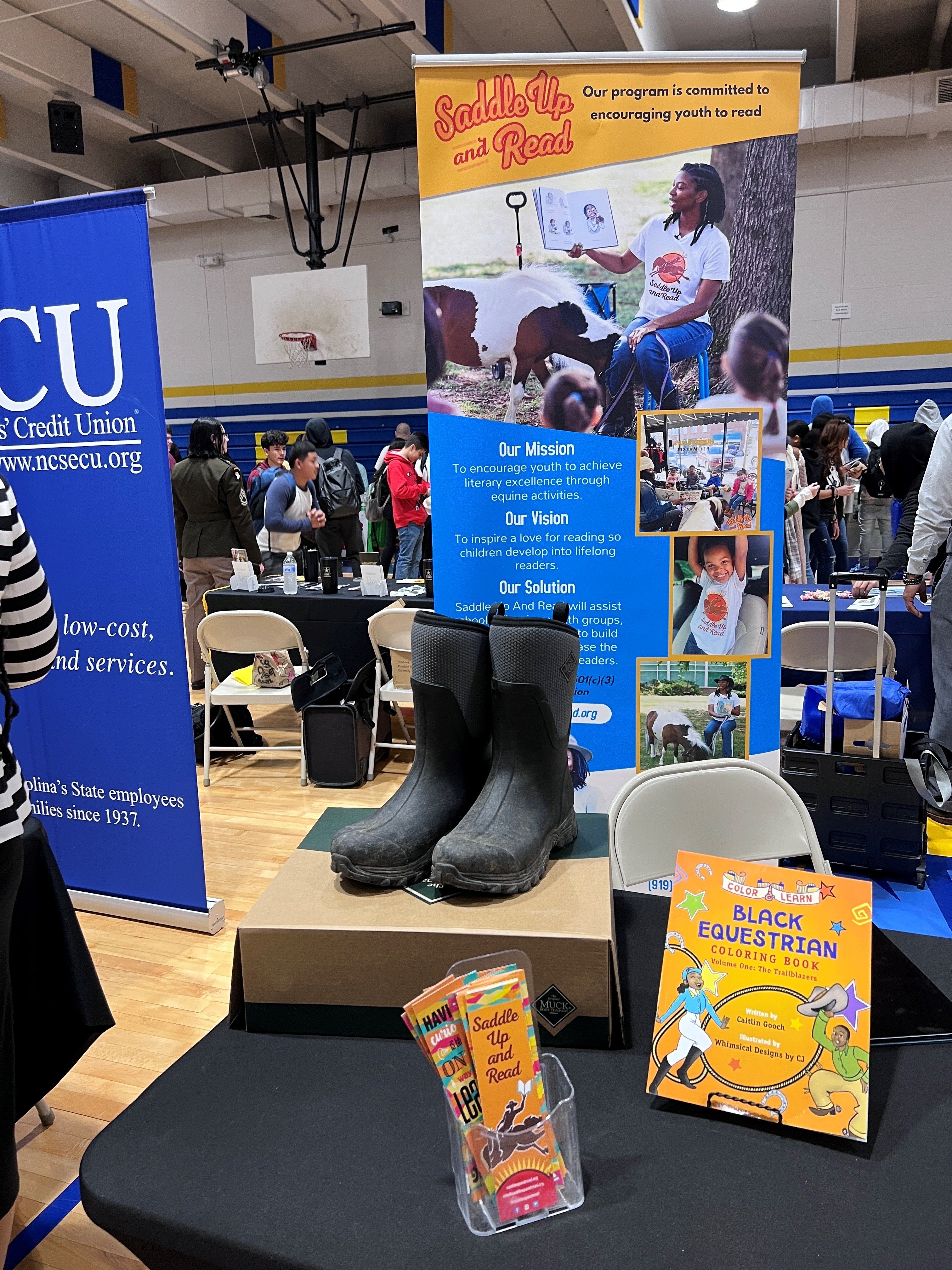 Promotional display for the Saddle Up and Read Program, inside the school gymnasium. Muck Boots posed on a table.